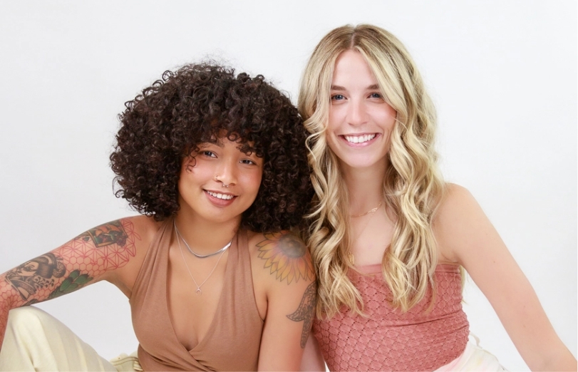 Two young women sitting next to each other. One has curly hair and the other has wavy blonde hair. This salon can accommodate any hair type.