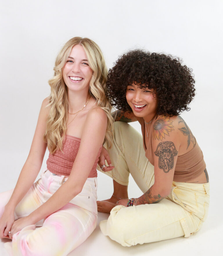 Two young women sitting next to each other. One has curly hair and the other has wavy blonde hair. This salon can accommodate any hair type.